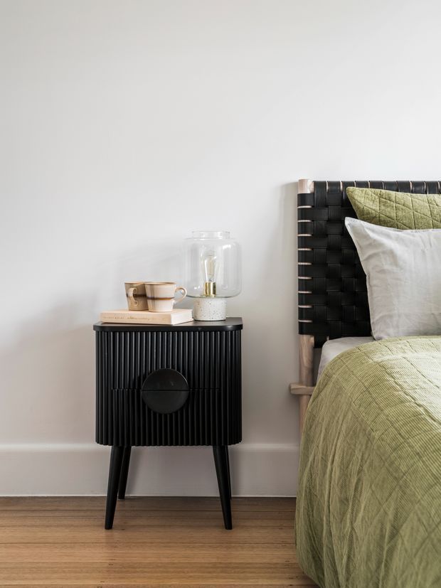 A close-up bedroom vignette featuring a black ribbed bedside table with tapered legs beside a bed. The bed has a woven black headboard, white and sage-green pillows, and a textured sage-green quilt. A ceramic cup, book, and glass vessel sit on the bedside table against a plain light wall, with warm timber flooring below.
