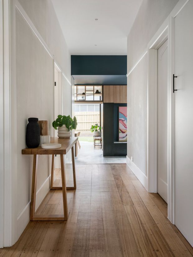 A bright, narrow residential hallway with white walls, simple trim, and a warm timber floor. A slim timber console table with ceramic vessels and a leafy plant sits along the left wall. The corridor leads toward an open living area with dark teal built-in joinery, timber cabinetry, glazing, and colourful artwork. White interior doors line the right side, and recessed ceiling lights provide understated illumination.