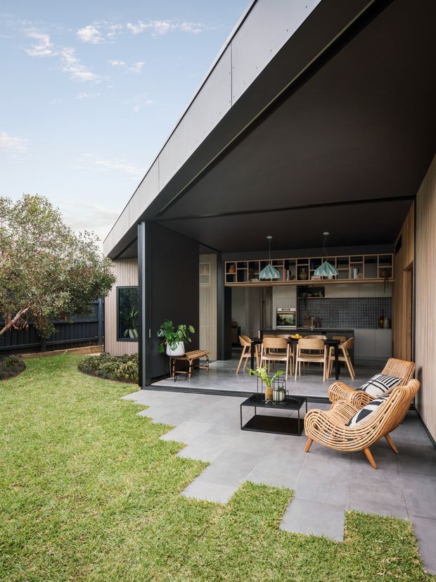 A contemporary indoor-outdoor living and dining area opening onto a landscaped lawn through large sliding glass doors. The covered terrace has grey tiled paving, wicker lounge chairs, a low black coffee table, and potted plants. Inside, a timber dining table with chairs sits beneath pendant lights, with dark cabinetry and a tiled backsplash. The home features a strong black roof and soffit line, vertical timber wall cladding, and a sheltered courtyard-like arrangement.