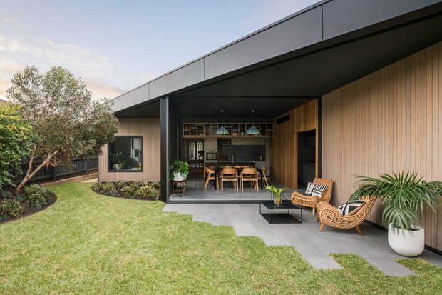 A low-slung contemporary home with a deep covered outdoor entertaining area opening onto a landscaped lawn. The facade combines warm vertical timber cladding with dark metal trim and a broad flat roof. A dining area and compact kitchen are visible beneath the overhang, while rattan lounge chairs, potted plants, and a paved terrace create a relaxed indoor-outdoor setting.