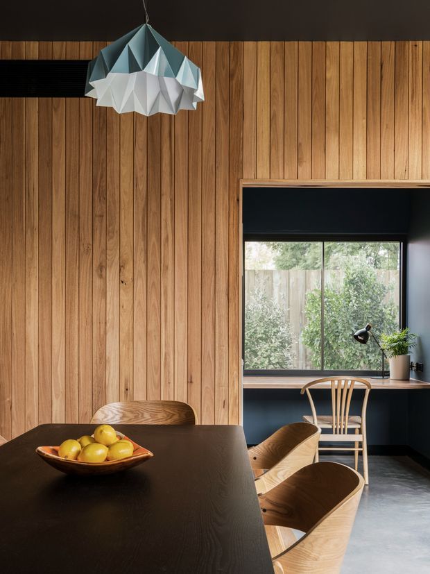 Warm, modern dining and study nook featuring full-height vertical timber wall panelling, a dark dining table with wooden chairs, and a compact built-in desk beneath a large window. A faceted geometric pendant light hangs above the table, while dark painted walls and a polished concrete floor add contrast to the natural timber.