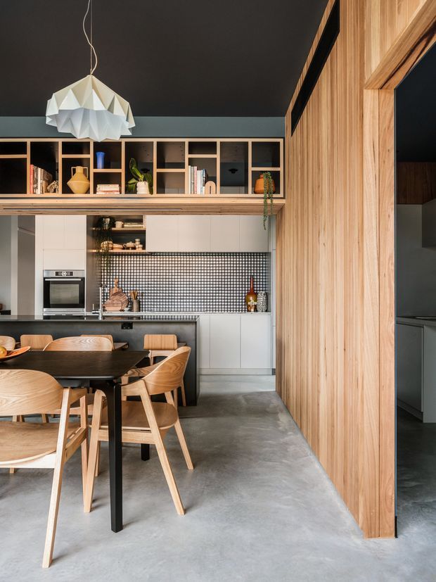 Contemporary open-plan kitchen and dining area with a dark ceiling, polished concrete floor, extensive light timber wall panelling and joinery, grey cabinetry, a small square-tile backsplash, open shelving, and a sculptural white pendant light. A dark dining table with timber chairs occupies the foreground.