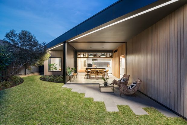 Exterior view of a contemporary single-storey home opening onto a covered outdoor entertaining area and landscaped lawn. The dark flat roof, broad cantilevered eaves, timber-clad side wall, large glazed opening, and warm integrated lighting frame a visible kitchen and dining area. Outdoor lounge chairs and a coffee table sit on a concrete-paved terrace.