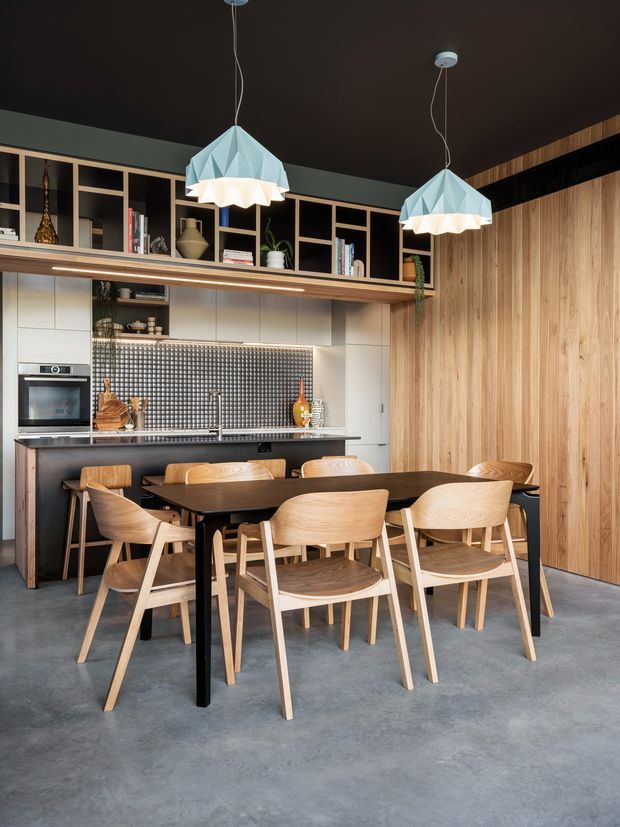 A contemporary open-plan kitchen and dining area with a dark ceiling, polished concrete floor, timber dining table and chairs, white built-in kitchen cabinetry, black island bench, and a textured dark mosaic tile splashback. Full-height vertical timber wall panelling and open timber shelving add warmth, while geometric pendant lights provide a decorative focal point.