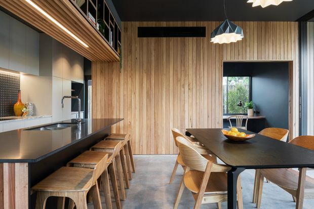 A sophisticated open-plan kitchen and dining area with a long dark island benchtop, timber bar stools, integrated cabinetry, vertical timber wall panelling, and a matching dark dining table with timber chairs. Black-framed glazing, a concrete-look floor, open shelving, and sculptural pendant lights create a warm contemporary interior.