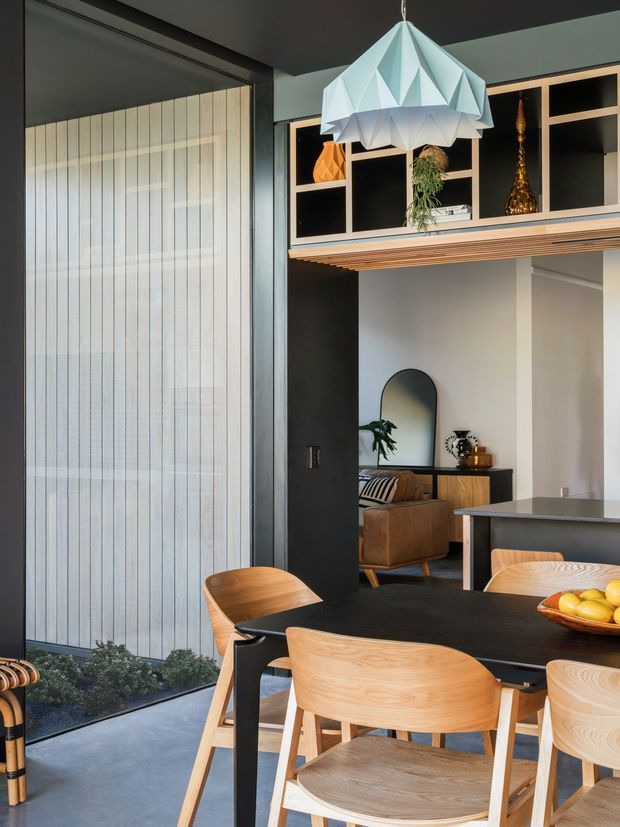 A refined open-plan dining and kitchen area with a black dining table surrounded by light timber chairs, a sculptural faceted pendant light, and built-in shelving with decorative objects. A living room with a sofa and arched mirror is visible beyond the opening. The space combines dark painted surfaces, warm timber joinery, and pale concrete-look flooring, with large translucent vertical blinds or screening at the glazed opening.