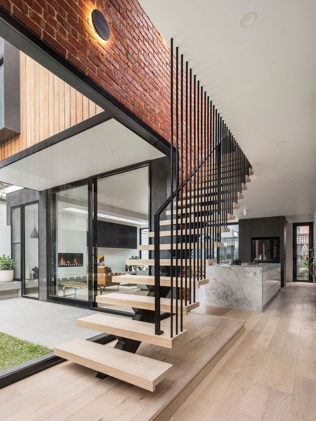 Contemporary open-plan interior featuring a floating timber staircase with slim black metal balustrade, exposed red-brick feature wall, expansive black-framed glazing, and a marble kitchen island. The space includes a living area with fireplace visible beyond the glass, pale timber flooring, and strong indoor-outdoor connections.