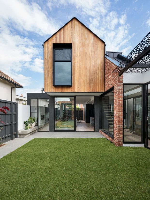 Exterior view of a contemporary multi-level residential extension arranged around a landscaped courtyard. The upper storey has a steep gable form clad in vertical natural timber boards, while the lower level combines extensive dark-framed glazing with an existing red-brick wall. A perforated metal canopy projects over the right side, and a lawn and paved courtyard occupy the foreground.