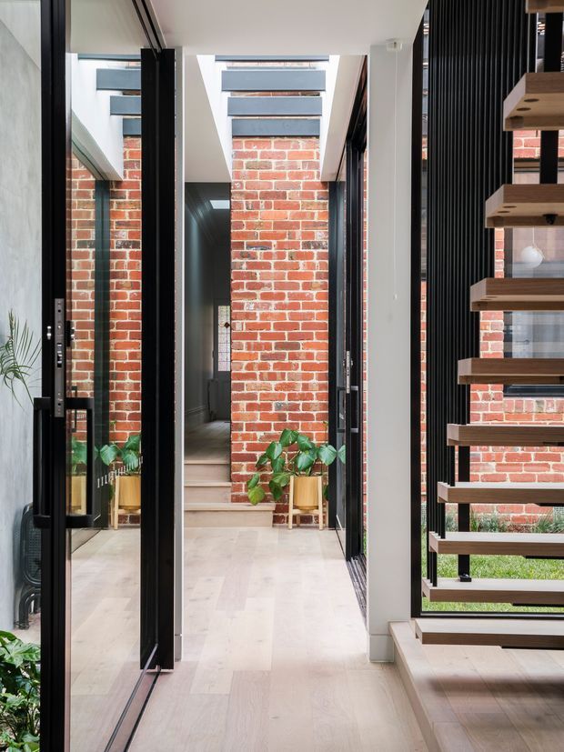 Bright contemporary interior hallway with pale timber flooring, exposed red brick walls, black-framed glazing and mirrored panels. A dramatic open-riser timber staircase with black vertical balustrade occupies the right side, with indoor plants adding greenery. The corridor leads toward a darker entry or adjoining space and is illuminated by skylights and large windows.