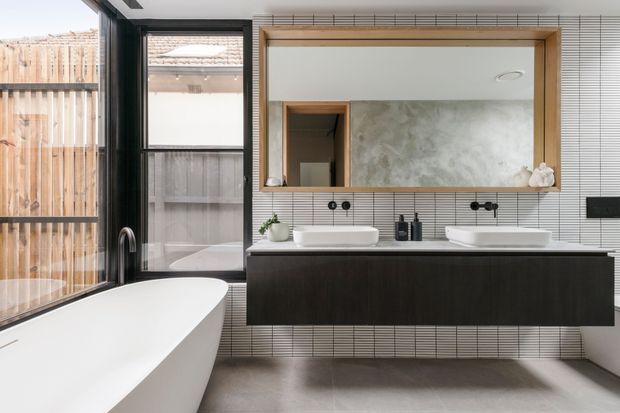 Contemporary bathroom featuring a freestanding white bathtub, double white vessel basins, a dark floating vanity, vertically lined light wall tiles, a large timber-framed mirror, black fixtures, and floor-to-ceiling glazing overlooking a timber-fenced outdoor area.