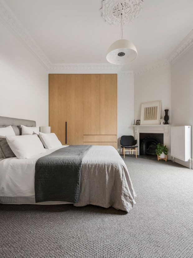 A bright, refined bedroom with a large upholstered bed dressed in white and grey linens, full-height light timber built-in wardrobe cabinetry, a traditional white fireplace with ornate detailing, grey carpet, and decorative plaster cornices and ceiling rose. A contemporary pendant light and simple black chair add contrast to the otherwise soft neutral scheme.
