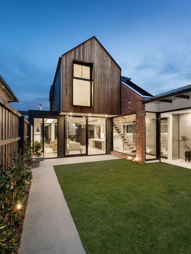 A dusk exterior view of a contemporary multi-level courtyard house. The building combines vertical weathered timber cladding, exposed red brick walls, black-framed floor-to-ceiling glazing, and a steep gabled upper volume. A central lawn and concrete path form the landscaped courtyard, with bright interior spaces visible through the glazing.