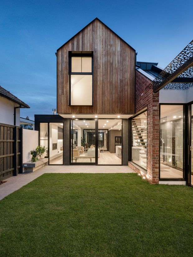 A dusk exterior view of a contemporary multi-level residential extension arranged around a small grass courtyard. The main gabled volume is clad in vertical weathered timber, with large dark-framed glazed doors and windows opening to the courtyard. An adjoining section features exposed red brick, a glazed stairwell, and a perforated overhead shade structure. Warm interior lighting is visible through the extensive glazing.