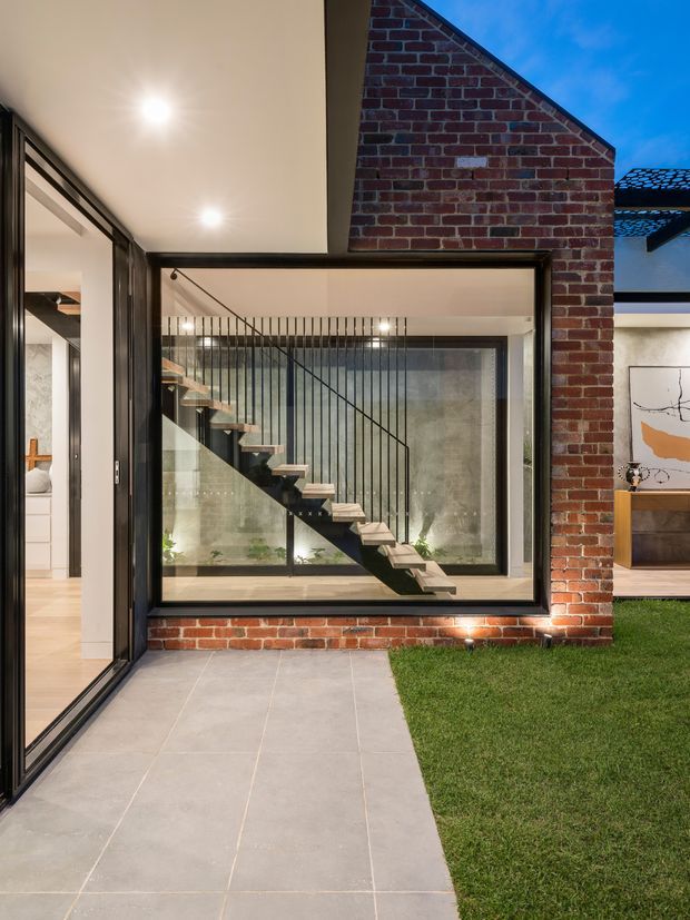 View from a paved courtyard or terrace into a contemporary house, centred on a dramatic floating staircase behind expansive black-framed glazing. The composition combines exposed red brick walls, concrete-look surfaces, timber stair treads, black metal balustrading and a small lawn, with interior living spaces visible through the windows and open glass door.