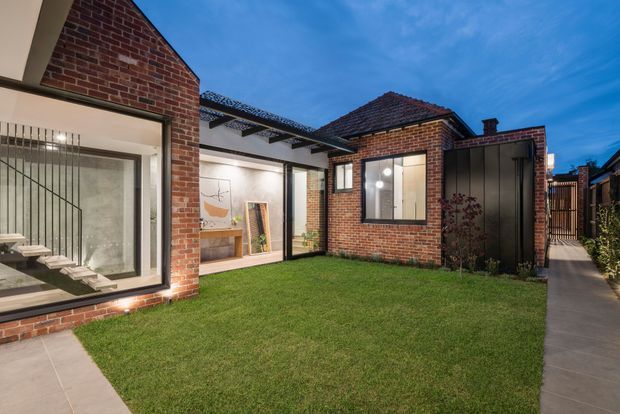 Twilight view of a contemporary residential courtyard framed by red brick buildings. The courtyard includes a manicured lawn, concrete paths, large glazed openings, a projecting floating staircase visible through the left window, and a covered outdoor connection with exposed dark beams. The composition combines retained brickwork with modern metal and glass elements.