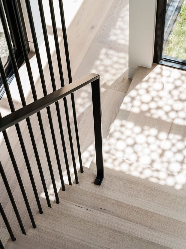 Close-up view of a modern stairway with pale timber treads and landing, slim black metal vertical balusters, and a matching black handrail. Large glazing beside the stairs casts patterned sunlight and shadows across the timber flooring.
