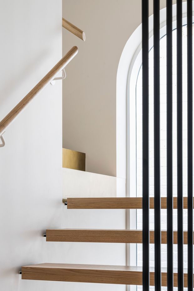 Close-up architectural view of a contemporary staircase with floating timber treads and a matching timber handrail against white walls. Black vertical metal balustrade bars run alongside a tall arched window, creating a strong contrast and allowing abundant natural light into the stairwell.