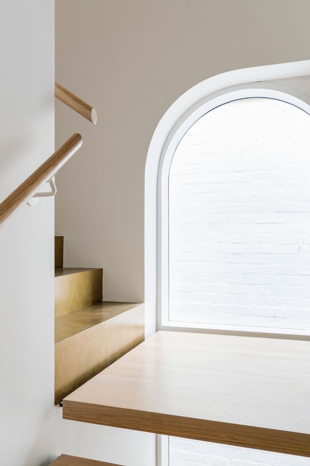 A bright, minimalist stairway with floating timber steps and matching cylindrical handrails, set against warm beige walls. The staircase meets a large arched window with a white frame, revealing a white-painted brick wall beyond. A light timber landing or platform occupies the foreground, with brass-toned stair surfaces adding a subtle metallic accent.