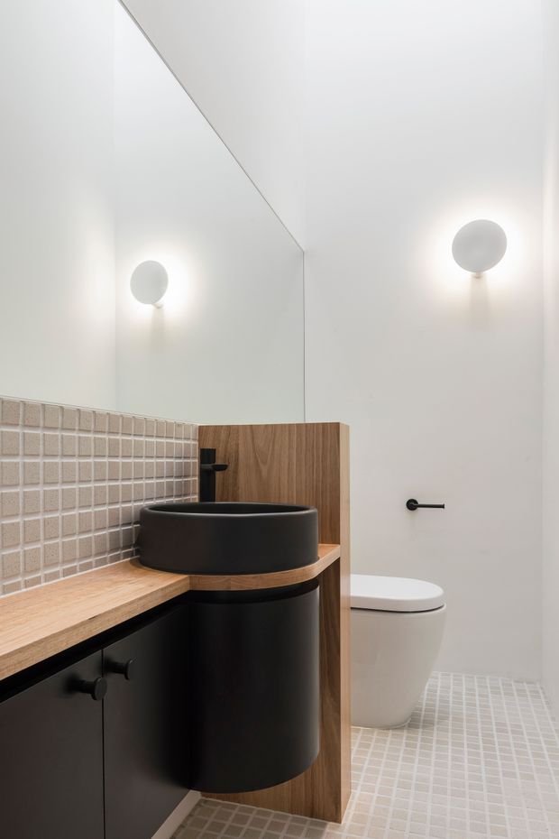 A compact contemporary bathroom with a floating timber vanity, matte black cylindrical vessel sink, black cabinetry and fixtures, pale beige square mosaic tile backsplash and floor, white walls, a wall-mounted round light, and a partially screened white toilet.