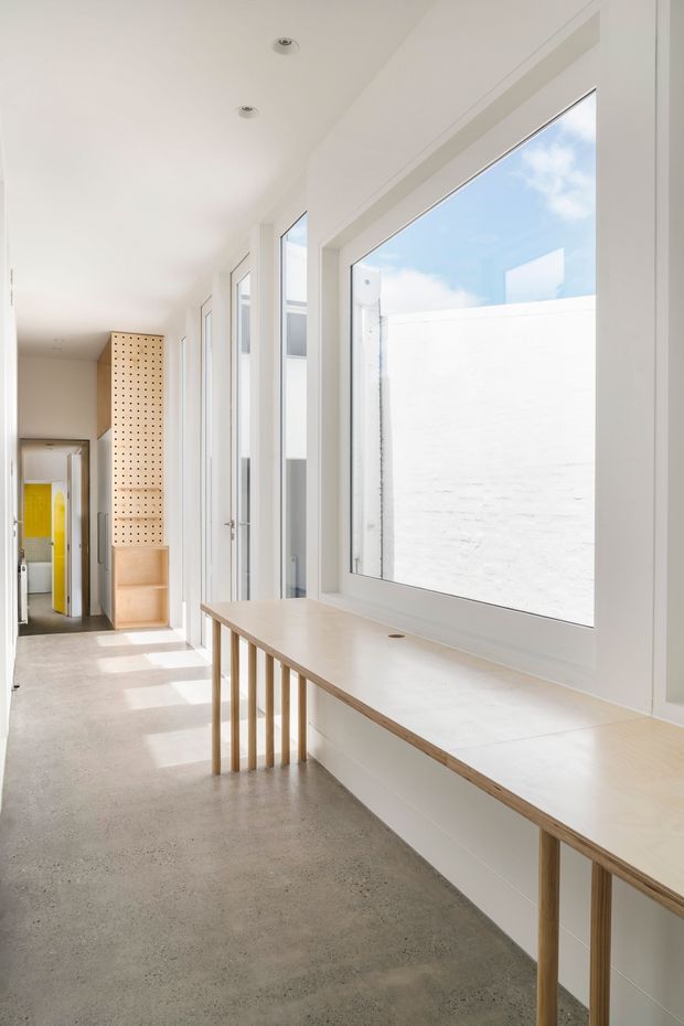 A bright, minimalist interior hallway or circulation space with a long built-in timber window seat and desk-like ledge beneath large white-framed windows. The space features polished concrete flooring, white walls and ceiling, recessed downlights, and a perforated timber screen with open shelving at the far end. A bathroom or utility area is partially visible through the doorway.