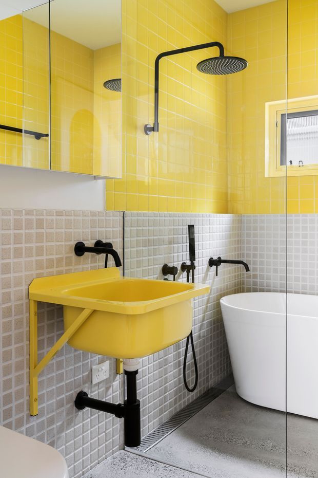 A compact, boldly coloured bathroom featuring bright yellow glazed wall tiles, pale grey square wall tiles, a vivid yellow wall-mounted basin with exposed black plumbing, matte black tapware and shower fittings, a white freestanding bathtub, mirrored cabinetry, and a small frosted window. The design has a playful graphic quality with strong yellow, white, grey, and black contrast.