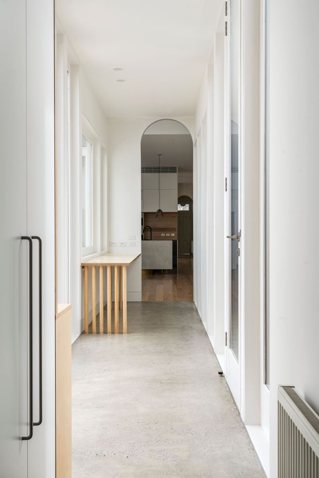 Bright minimalist residential hallway with white walls, tall windows and doors, a pale polished concrete floor, and a timber stair balustrade. The corridor leads through an arched opening to a contemporary kitchen with white cabinetry, a dark benchtop, pendant lighting, and timber flooring.