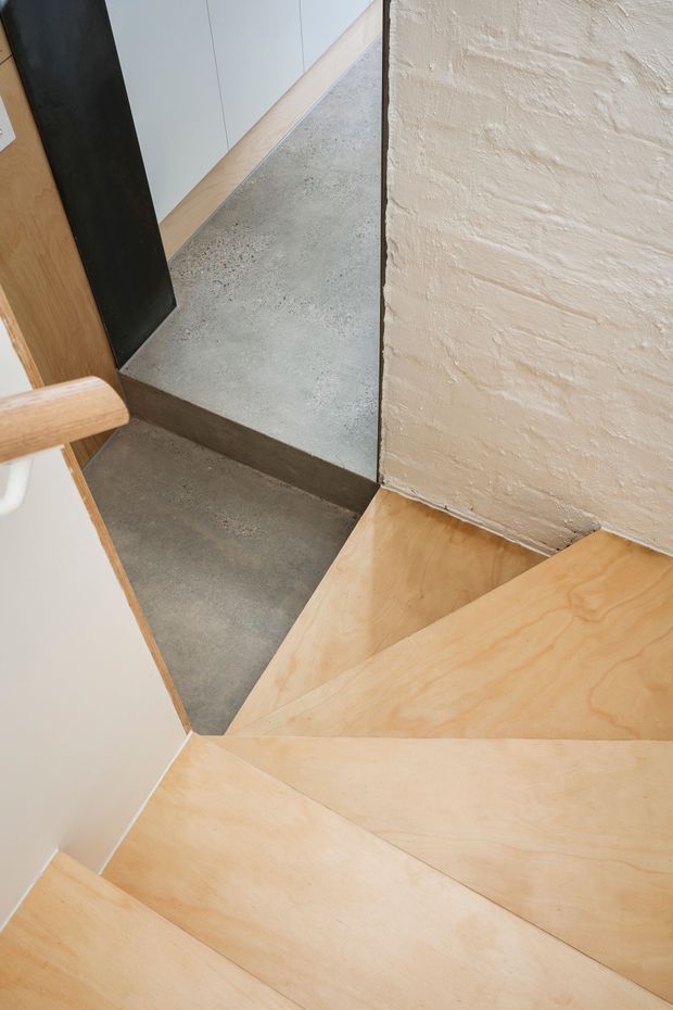 Close-up architectural photograph looking down a compact stairway with light timber treads, a dark concrete landing, a black vertical metal element, and a textured off-white plaster wall. The composition highlights the junction of contrasting materials and the stair geometry.