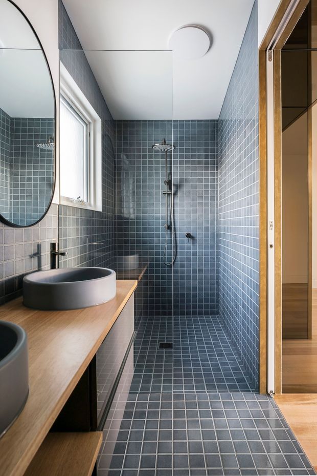 A compact modern bathroom with a walk-in tiled shower, wall-mounted shower rail, round vessel basins on a timber vanity, and a large rounded mirror. Small blue-grey square tiles cover the shower walls and floor, contrasted with warm timber cabinetry and door framing. A frosted window provides natural light, while a simple circular ceiling light adds a clean contemporary detail.