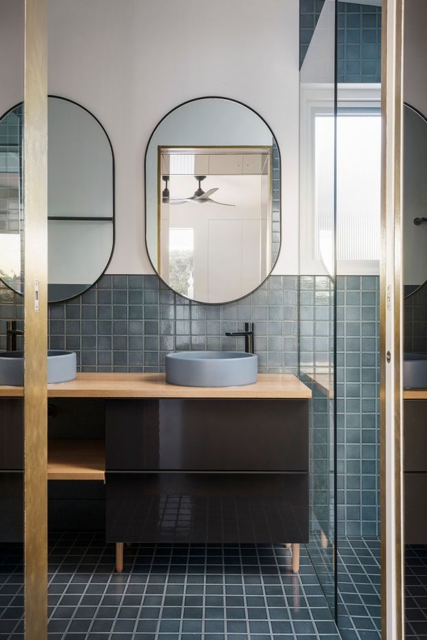 A contemporary bathroom vanity with a round vessel basin, wall-mounted black cabinetry, timber benchtop, and dark blue-grey square tiled walls and floor. Arched black-framed mirrors, a glass shower enclosure, and warm timber-framed sliding doors add strong geometric and material contrast. A bedroom or adjacent room is visible through the central mirror reflection.
