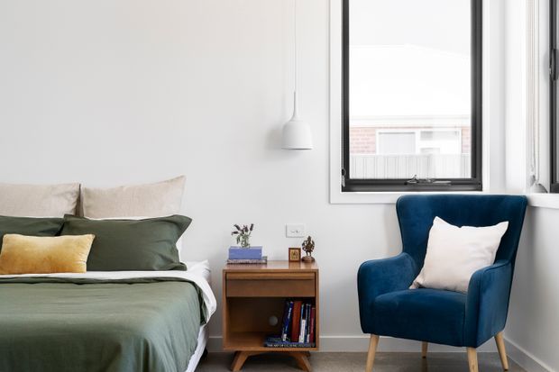 A bright, compact bedroom with white walls, a dark green bedspread, layered neutral and green pillows, and a navy-blue upholstered armchair. A mid-century-style timber bedside cabinet with books and small decor sits beside the bed, with a simple white pendant light and black-framed window completing the room.