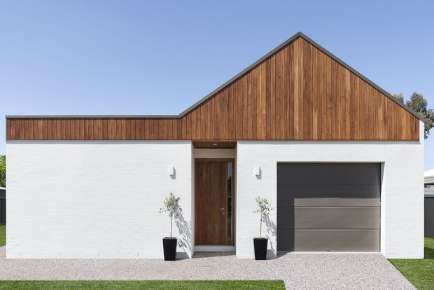 Front exterior of a contemporary single-storey home with a simple gabled roof form. The facade combines white rendered masonry walls with warm vertical timber cladding in the gable and parapet-like upper sections. A recessed timber entry door and dark sectional garage door sit beneath the roofline, with minimal landscaping and a gravel/concrete pathway.