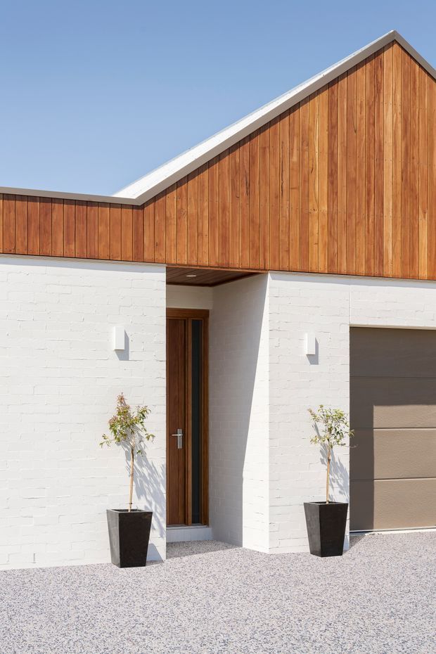 A bright contemporary residential facade with white-painted brick walls, warm vertical timber cladding beneath a prominent gable roof, a recessed timber entry door, and an integrated garage. Minimal wall-mounted lights and two potted trees frame the entrance, with a pale gravel forecourt in front.