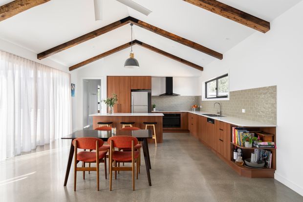 Bright open-plan kitchen and dining area with warm timber cabinetry, a long island bench, integrated appliances, textured light tile splashback, polished concrete flooring, and exposed dark timber ceiling beams beneath a pitched ceiling. A dining table and chairs occupy the foreground, with full-height sheer curtains and large windows bringing in natural light.
