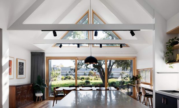 Bright contemporary open-plan kitchen and dining space with a dramatic pitched, raftered ceiling and clerestory glazing. Large timber-framed windows and doors open the room to a landscaped garden, while black linear track lighting, a reflective stone island, dark lower cabinetry, timber flooring, and built-in shelving create a refined modern interior. A dining table and chairs sit centrally, with a compact kitchen visible at right.