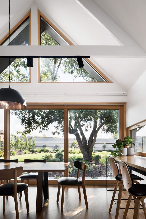 Bright, double-height dining and work area with extensive timber-framed glazing overlooking a landscaped garden. A pitched, angular white ceiling incorporates triangular clerestory windows and a linear black pendant light. The room contains a large timber dining table with upholstered chairs, plus a built-in timber desk or counter along the right wall. The space feels airy, warm, and contemporary with strong indoor-outdoor connection.