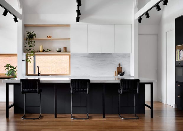Contemporary kitchen with a large black island and three black bar stools, white flat-panel cabinetry, and a pale marble-look splashback. Open timber shelving frames a window with exposed brick visible outside, while black track lighting and warm timber flooring add contrast. The room has a clean, refined, high-contrast look.