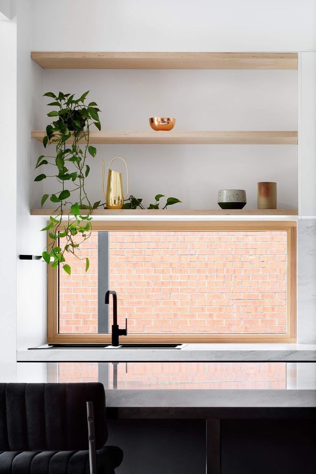 A bright, minimalist kitchen or kitchen workspace with pale timber open shelving, a timber-framed horizontal window, and a black tap and sink set into a light benchtop. The shelves display a trailing indoor plant and small decorative vessels. A dark dining or workspace table and upholstered chair occupy the foreground, while a red brick wall is visible through the window.