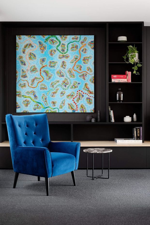 A refined seating nook featuring a vivid cobalt-blue tufted armchair, a small round marble-top side table, and a dark built-in wall of cabinetry and open shelving. The joinery incorporates a large colourful abstract artwork, books, ceramics, and a trailing plant, with a pale timber ledge and grey carpet completing the interior.