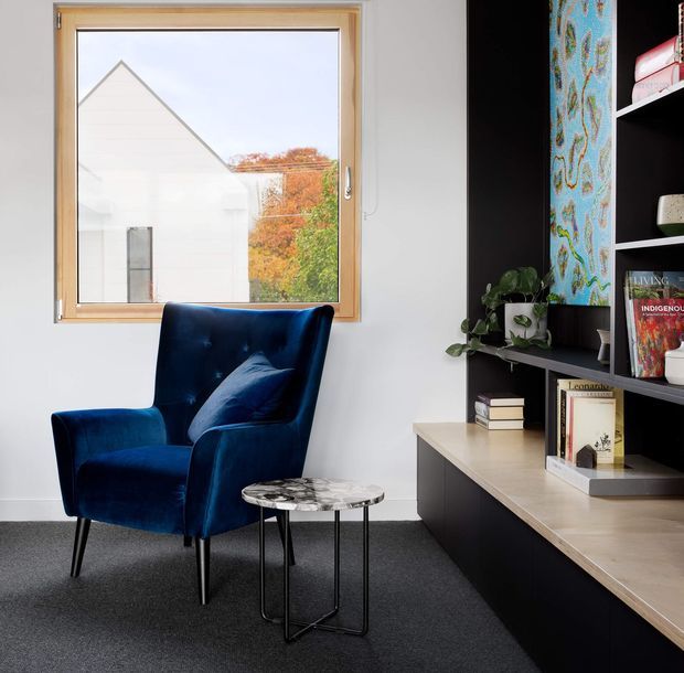 A bright, compact reading nook or study corner with a deep navy-blue upholstered wingback armchair, a small round marble-look side table, and a built-in black shelving and cabinet unit with a light timber benchtop. A large timber-framed window provides daylight and overlooks neighbouring architecture and autumn foliage. The built-in shelving includes books, plants, and a colourful decorative vertical panel.