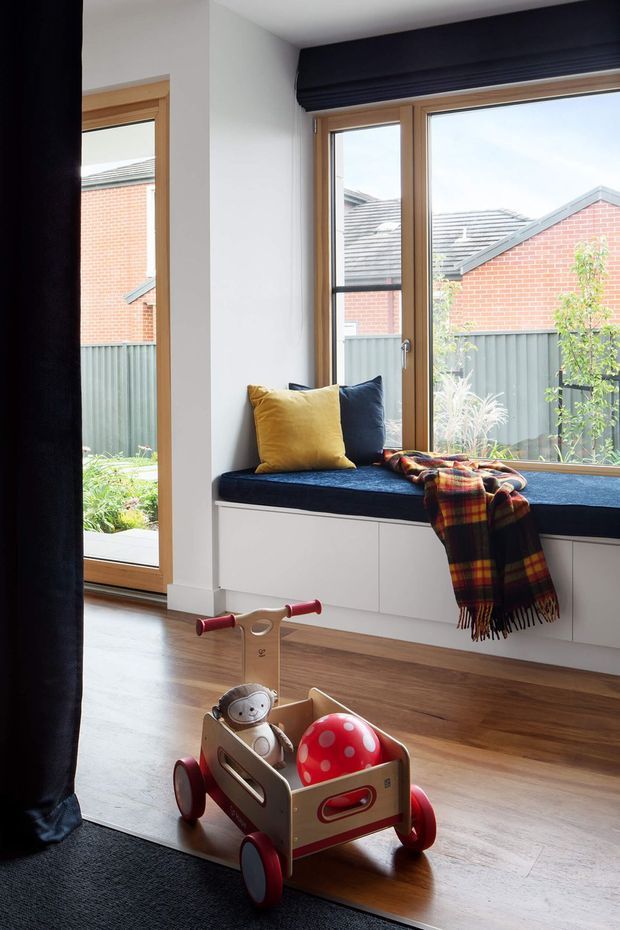 Bright, cozy interior corner featuring a built-in window seat beneath large timber-framed windows. The seat has dark blue upholstery, white built-in cabinetry below, mustard and navy cushions, and a plaid throw. Warm timber flooring, dark curtains, and a child's wooden push wagon create a comfortable family-oriented atmosphere. A fenced garden and neighbouring brick buildings are visible outside.