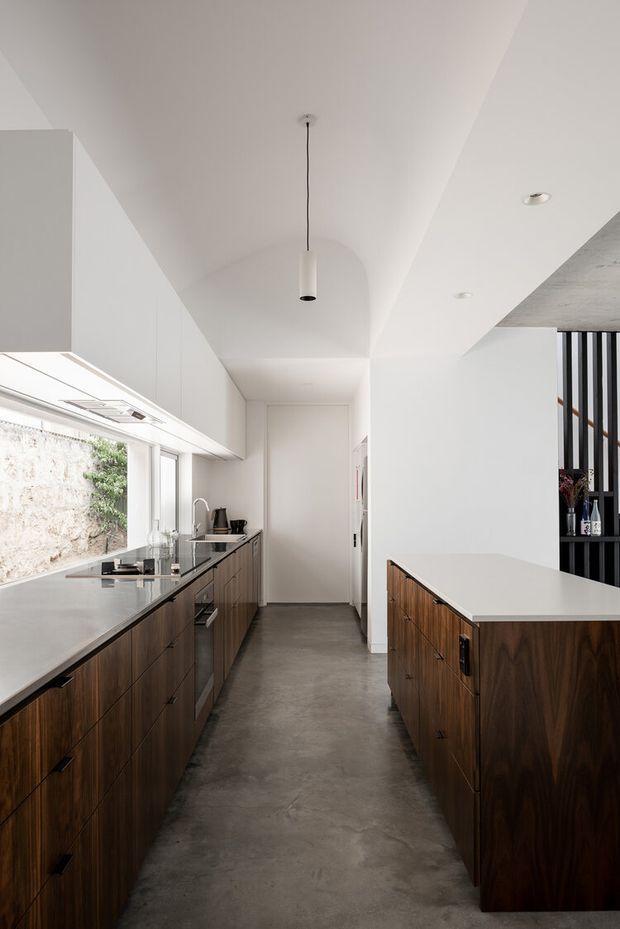 A narrow, contemporary galley kitchen with extensive dark timber cabinetry on both sides, pale solid-surface benchtops, white overhead cabinetry, and a polished concrete floor. A large horizontal window brings in natural light and overlooks a textured exterior wall with greenery. A central pendant light, recessed ceiling lights, and a black vertical screen with open shelving add restrained architectural detail.