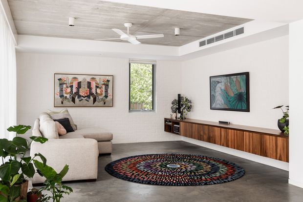 Bright contemporary living room with a large L-shaped neutral sofa, dark circular patterned rug, floating timber entertainment joinery, framed artwork, indoor plants, and a ceiling fan. The room features white painted brick walls, polished concrete flooring, and a recessed concrete-look ceiling panel with integrated ventilation.