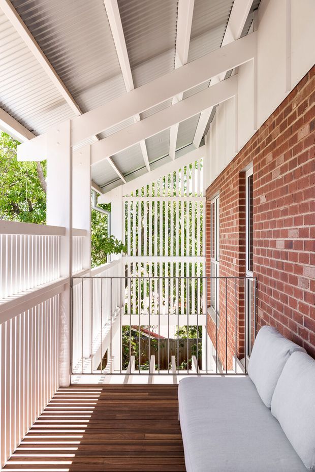 A bright covered outdoor balcony or terrace with a dark timber deck, white vertical screening and balustrades, exposed white-painted roof beams, and a red brick wall. A light grey outdoor sofa occupies the right side, while leafy greenery is visible beyond the slatted screen. Strong linear shadows create a crisp, airy architectural feel.
