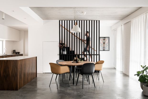 Bright open-plan interior showing a dining area beside a compact kitchen. A dark timber island and cabinetry sit to the left, while a round dining table with mixed tan and black chairs occupies the centre. A black vertical-slat screen partially conceals a stairway, with a person visible ascending behind it. The space has a polished grey concrete floor, a pale concrete-look ceiling, white walls, large sheer-curtained glazing, and minimalist pendant lighting.