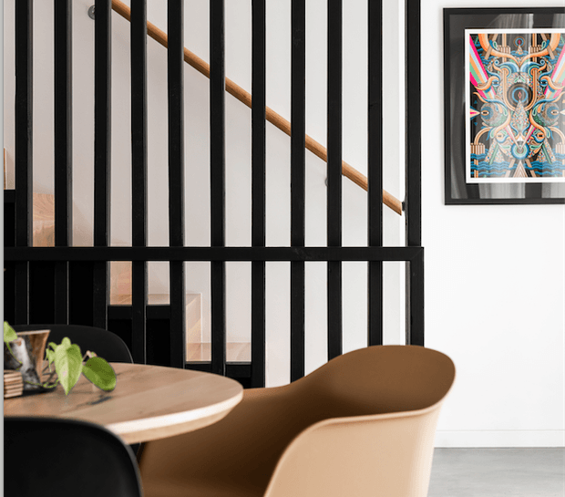 A contemporary dining area beside a black vertical-slat stair balustrade. The scene features a round light-wood dining table, sculptural tan dining chairs, and a black chair in the foreground. A timber handrail and staircase are visible behind the slatted divider, with colourful framed artwork on a white wall and a pale grey floor.