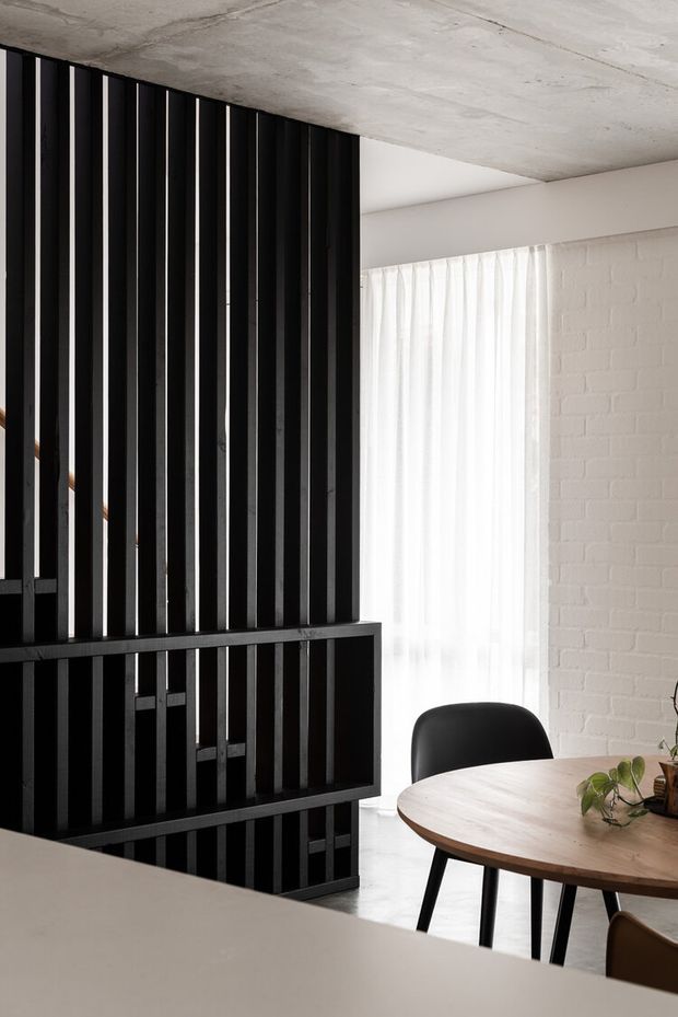A refined dining area with a round timber dining table and black chairs, positioned beside a sheer-curtained window and white-painted brick wall. A dramatic black vertical-slat screen and balustrade divide the space, while the exposed concrete ceiling and restrained monochrome palette create a modern industrial character.