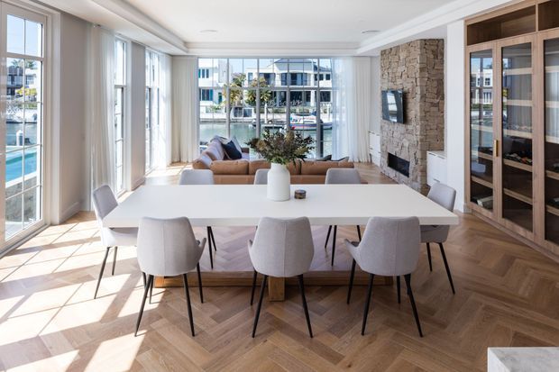 Bright, spacious open-plan dining and living area with floor-to-ceiling glazing, a large white dining table, upholstered dining chairs, and a pale sofa seating area. The room features herringbone timber flooring, white walls and ceiling detailing, extensive built-in timber cabinetry, and a stacked-stone fireplace wall. Waterfront buildings and water are visible beyond the glazing.
