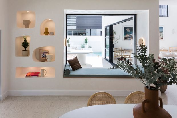 Bright contemporary dining area with a large black-framed interior window or pass-through opening overlooking a light-filled courtyard and adjoining seating area. The wall features recessed display niches with integrated warm lighting, pottery and small decorative objects, while a round dining table, woven-back chairs and a large leafy arrangement occupy the foreground. Pale walls and terrazzo-like flooring create a clean, warm, refined atmosphere.