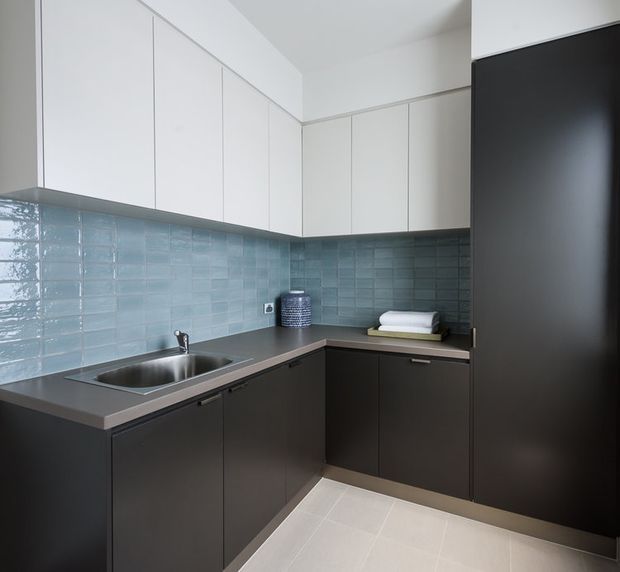 Compact contemporary galley kitchen with L-shaped dark lower cabinetry, white handleless overhead cupboards, a stainless-steel sink and benchtop, pale blue-green glazed rectangular tile splashback, and light tiled flooring. A tall dark integrated appliance or storage unit occupies the right side.
