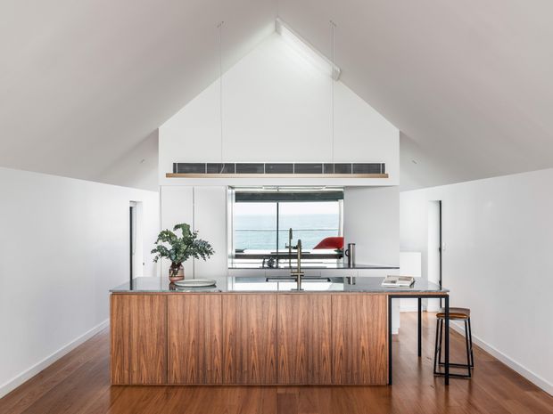 Bright contemporary kitchen in a compact open-plan space with a steep vaulted ceiling and white walls. A timber-clad island with a pale stone benchtop dominates the foreground, paired with black-framed stools and a dark linear rangehood or shelf feature. A large window behind the sink overlooks a coastal or waterfront scene, while warm timber flooring and minimal furnishings create a clean, airy interior.